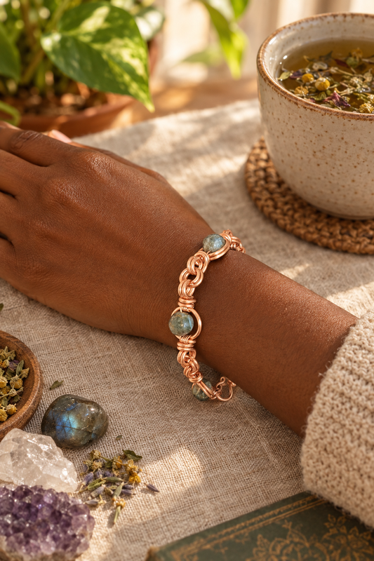 Copper-wrapped labradorite bracelet worn on a woman's wrist beside a cup of herbal tea and crystals